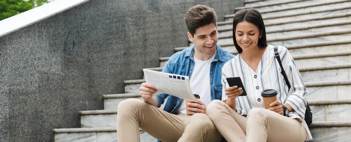 Junges Paar sitzt auf einer Treppe, sie liest am Smartphone, er hält eine Zeitung; beide trinken Kaffee und lächeln.